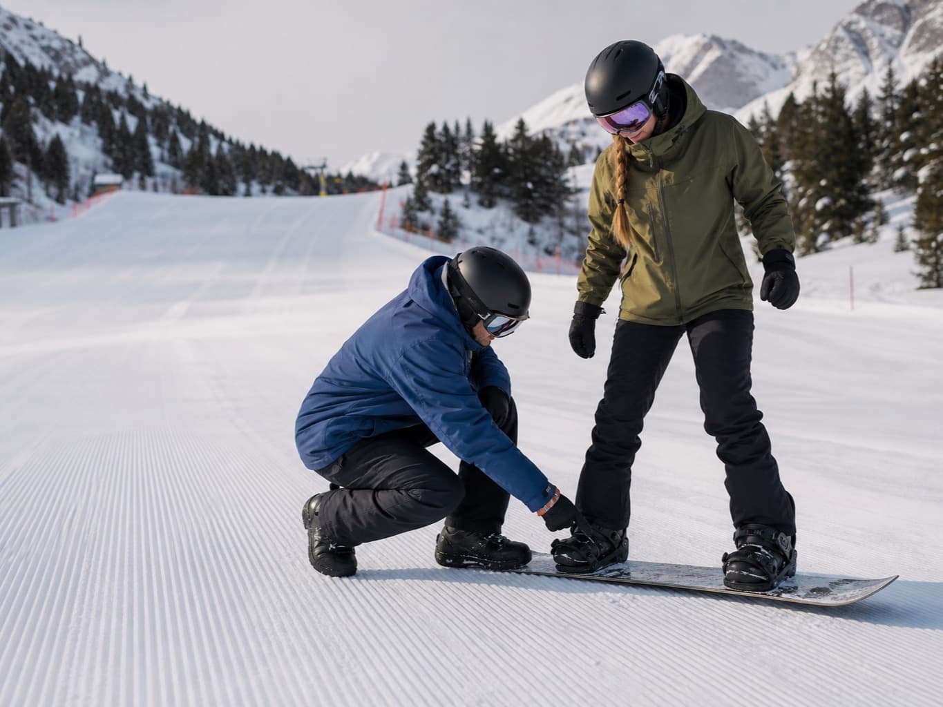 Javi crouched beside a rider, fixing their stance on the board on freshly groomed piste